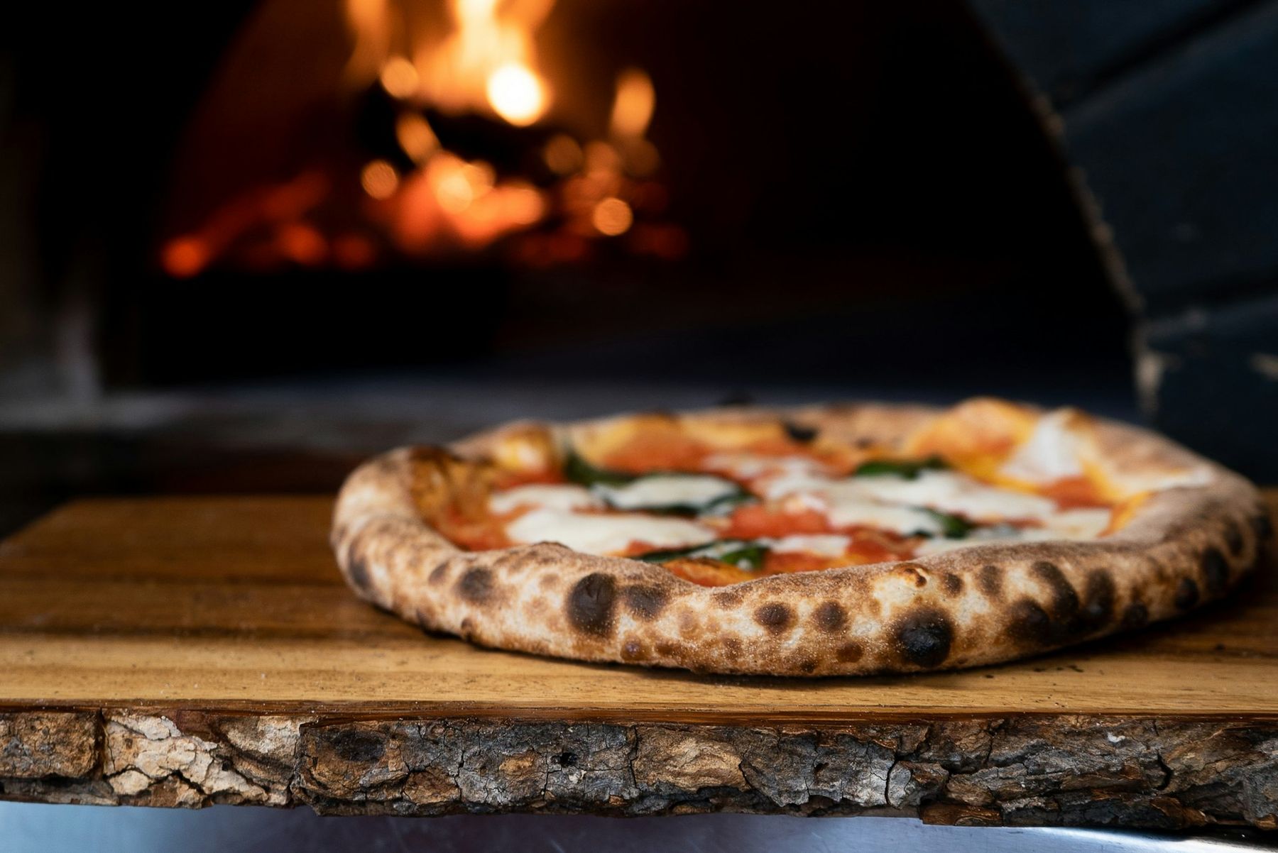 Pizza appena sfornata su tavola di legno davanti al forno a legna acceso